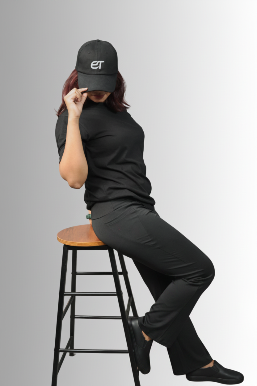 A woman wearing a black t-shirt, black pants, and a black baseball cap with a white 'et' logo sits on a wooden-topped stool against a light gray background.